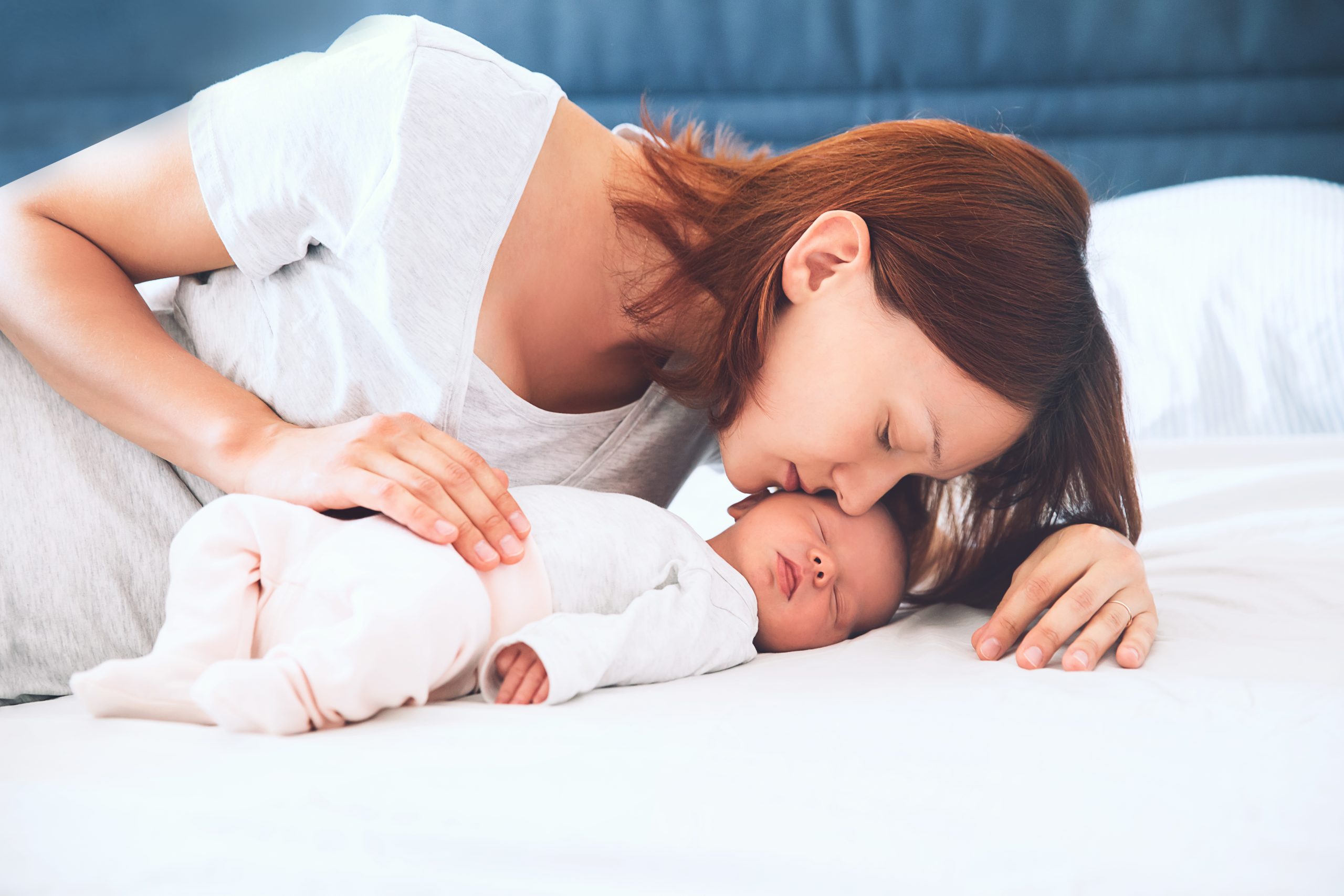 Mother kissing her newborn baby. Young beautiful mom lying at bed with a cute little sleeping child. New born baby's first days of life in family at home. Loving mother looks at asleep infant.